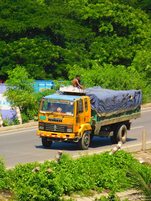 A yellow flatbed truck parked on a residential street during daylight hours, with a grey tarpaulin covering its cargo area. A person is sitting on top of the truck, which is positioned near a curb and surrounded by lush green trees and bushes. The street features a sidewalk with white and red striped traffic bollards along the edge, and the background includes dense foliage, suggesting a suburban or semi-urban environment. This scene illustrates the transportation phase of a home relocation, involving furniture or boxes being prepared for moving, with the vehicle ready for loading or unloading as part of a professional removals service by Man with Van Snaresbrook.