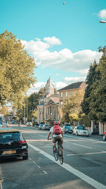 A cyclist wearing a red backpack and a white helmet rides along a paved street lined with parked cars on both sides, with trees displaying green foliage on the left and right. In the background, there are historic-style buildings made of stone or brick, some with large windows, and a prominent church with a tall clock tower featuring a pointed spire. The sky above is bright blue with scattered white clouds, providing natural lighting for the scene. The street appears to be part of a residential or mixed-use area in Snaresbrook, with additional vehicles visible further down the road. The image captures a quiet moment during daytime, involving urban transportation and local architecture, relevant to home relocation and moving logistics in the area.