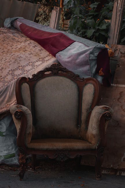 The image shows an antique wooden armchair with an upholstered fabric seat and backrest, positioned indoors on a wooden floor. Behind the armchair, there is a large piece of fabric-covered furniture, possibly a sofa or extended cushion, with a mix of grey, maroon, and other fabric sections draped over it. To the left, a plastic bag containing some items is visible, and the background includes a window or door frame leading to an outdoor garden with green foliage. The lighting appears natural, with daylight illuminating the scene, suggesting the setting may be a storage or staging area for house removals. The environment indicates packing and furniture transportation activities, with the visible furniture and materials likely prepared for moving or disposal, consistent with services provided by Man with Van Snaresbrook.