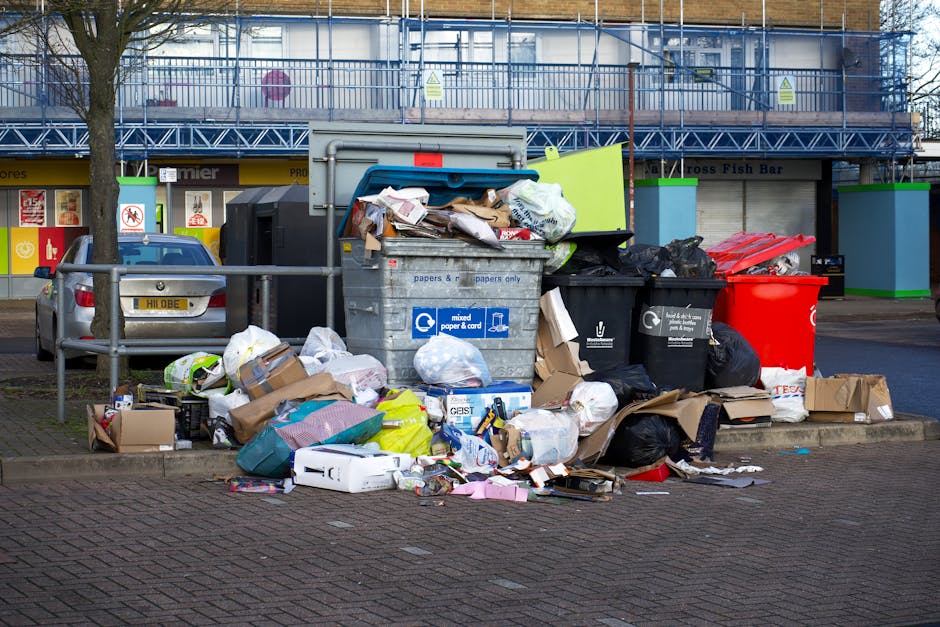 A collection of overflowing waste and recycling bins situated on a paved sidewalk in front of a building with a blue scaffold and signage for a fish bar. The waste includes various cardboard boxes, black plastic rubbish bags, and paper and packaging materials. Some cardboard boxes are flattened, while others are partially open, revealing contents. The bins include a large grey mixed waste container, a black recycling bin, a red waste bin, and a black bin with a white recycling symbol, all positioned on a curb with a metal railing nearby. The scene is illuminated by natural daylight, and the surrounding environment features parked cars and a tree, reflecting typical urban waste collection areas. The image highlights the need for efficient rubbish removal and clearance services, as offered by Man with Van Snaresbrook, for home relocations or waste disposal tasks.