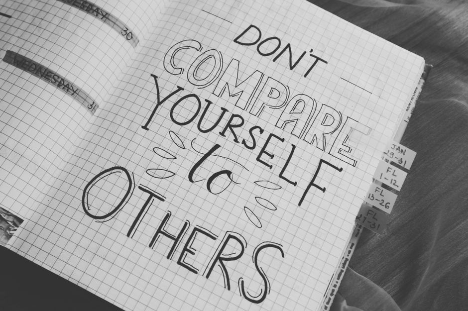 A hand-drawn note on a graph paper notebook page with the message 'Don't compare yourself to others' written in large, mixed font styles, with decorative lines around the words. The notebook is positioned on a wooden surface, with visible tab dividers on the right side labeled with dates and initials. The scene suggests an environment focused on planning or personal motivation, relevant to home relocation or packing activities associated with professional removals or moving services, such as those offered by Man with Van Snaresbrook.