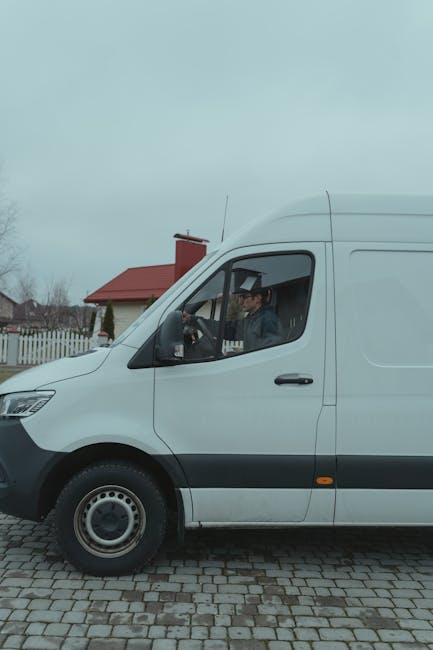 A white commercial van, part of a home relocation or furniture transport service, parked on a cobblestone driveway in front of a house with a red roof and white picket fence. The driver, visible through the window, is wearing a dark jacket and glasses, seated inside the vehicle. Inside the van, the side door is open, revealing the interior which is likely used for loading and unloading boxed items, furniture, and packing materials during a move. The scene is set in daylight, with overcast skies and residential surroundings visible in the background. The image reflects the vehicle used by [COMPANY_NAME] to assist with packing, moving, and logistics during house removals or related moving services in Snaresbrook.