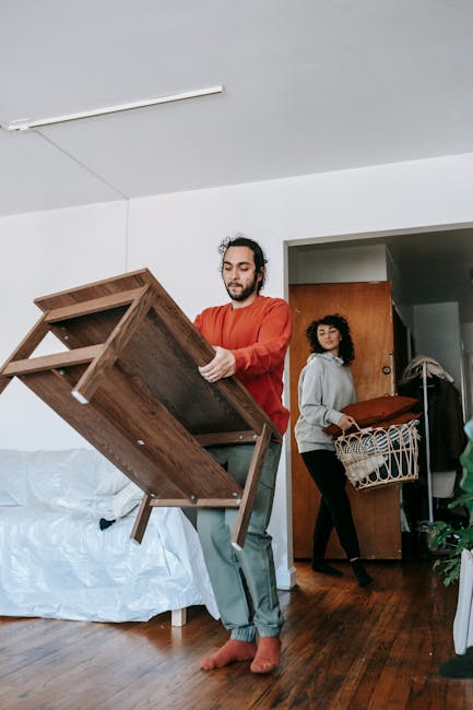 Inside a residential room during a home relocation, a small black and white cardboard box is placed on the wooden floor containing an incomplete white and black guitar; nearby, an open brown cardboard
