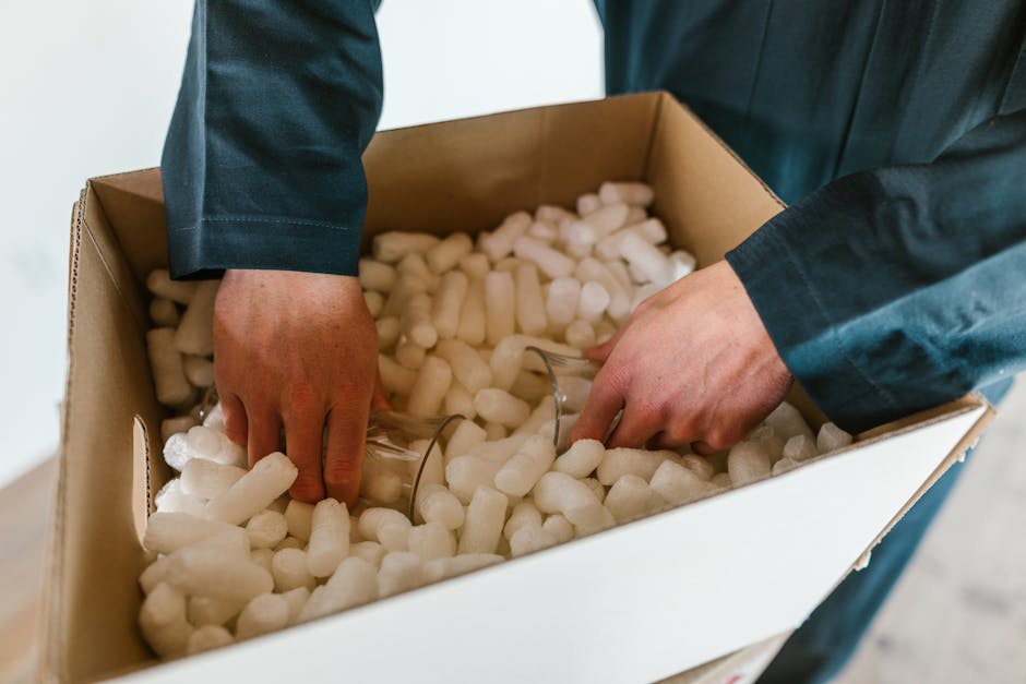 A person wearing a dark suit is placing a fragile glass item into a cardboard box filled with white packing peanuts for cushioning. The box is on a flat surface indoors, likely prepared for a home relocation or furniture transport process. The packing materials and careful handling suggest the preservation of delicate items during packing and moving managed by Man with Van Snaresbrook, supporting efficient packing and moving logistics for house removals in Snaresbrook. The scene captures the detailed action of packing fragile belongings for a household move, emphasizing the importance of secure packaging during furniture transport or packing and moving services within a home environment.