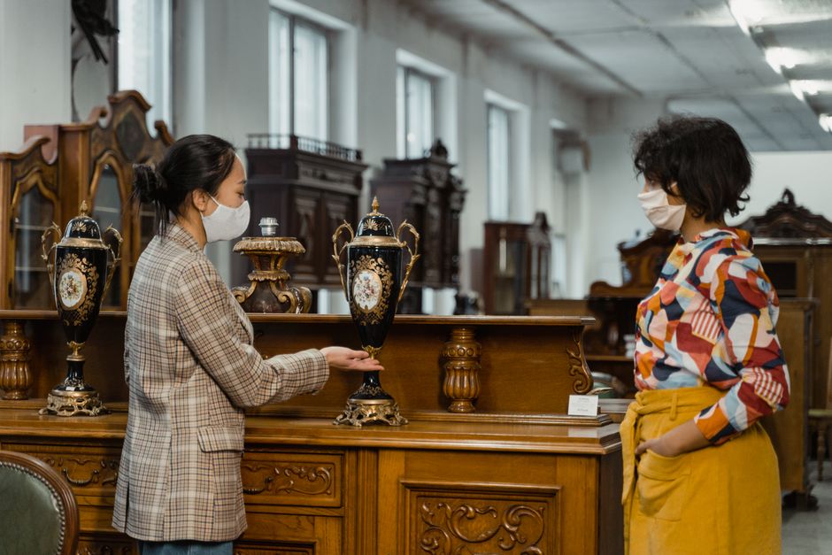 Two women wearing white face masks are inspecting decorative antique vases placed on a polished wooden table in an antique showroom. The woman on the left, dressed in a beige plaid blazer, is holding and examining one of the black and gold urn-shaped vases with floral patterns and ornate bases. The woman on the right, wearing a colorful patterned shirt and mustard yellow trousers, stands with her hands in her pockets, observing the items. The background features additional antique furniture and cabinets in a well-lit room with large windows, consistent with a house removal or furniture transport setting. The scene captures the careful handling and assessment of valuable items during a home relocation or antique moving process, supported by the professional environment of Man with Van Snaresbrook.