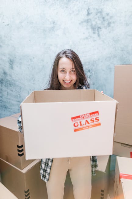 A young woman with long dark hair smiling while holding an empty cardboard moving box labeled 'Glass' in a well-lit indoor space with a light blue textured wall in the background. Surrounding her are 