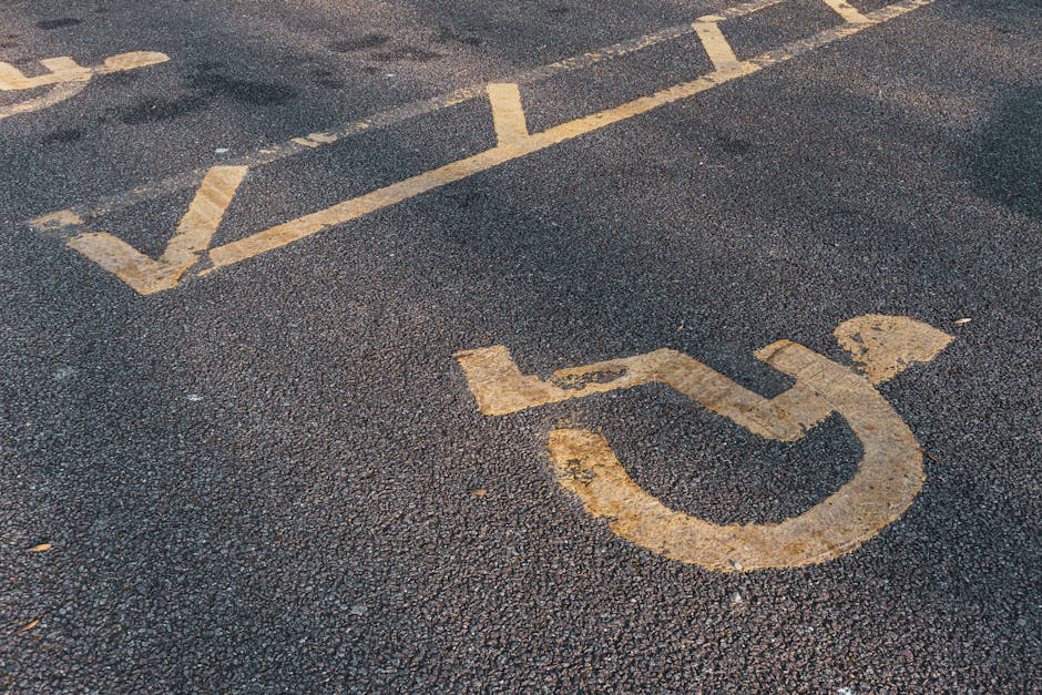 Close-up of a handicapped parking space symbol painted on an asphalt surface near the entrance of a property, with fading yellow lines and a worn wheelchair icon. The image shows the textured black asphalt with patches of lighter and darker areas, and the painted markings indicating designated parking for disabled individuals. The setting suggests the parking area is part of a home relocation or moving site, where a vehicle used by Man with Van Snaresbrook is parked nearby for furniture transport and loading. Visible around the edges are sections of the pavement, with no vehicles or furniture present in the frame, focusing solely on the parking space markings relevant to access and parking logistics for move-associated activities near Snaresbrook Crown Court.
