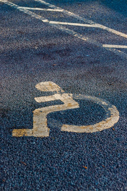 Close-up view of a public parking space featuring a white wheelchair symbol painted on dark asphalt surface, with visible wear and some fading. In the background, a partially visible white van is parked adjacent to the marked area, indicating the parking spot is designated for disabled access. The scene suggests an environment near a residential or commercial building, where home relocation services by Man with Van Snaresbrook may utilize this parking zone for loading or unloading furniture and household items during a house move. The image highlights the importance of accessible parking in the moving process, with emphasis on loading equipment such as trolleys or straps used to safely transfer packed boxes, furniture, and appliances from the property to the vehicle in an efficient manner.