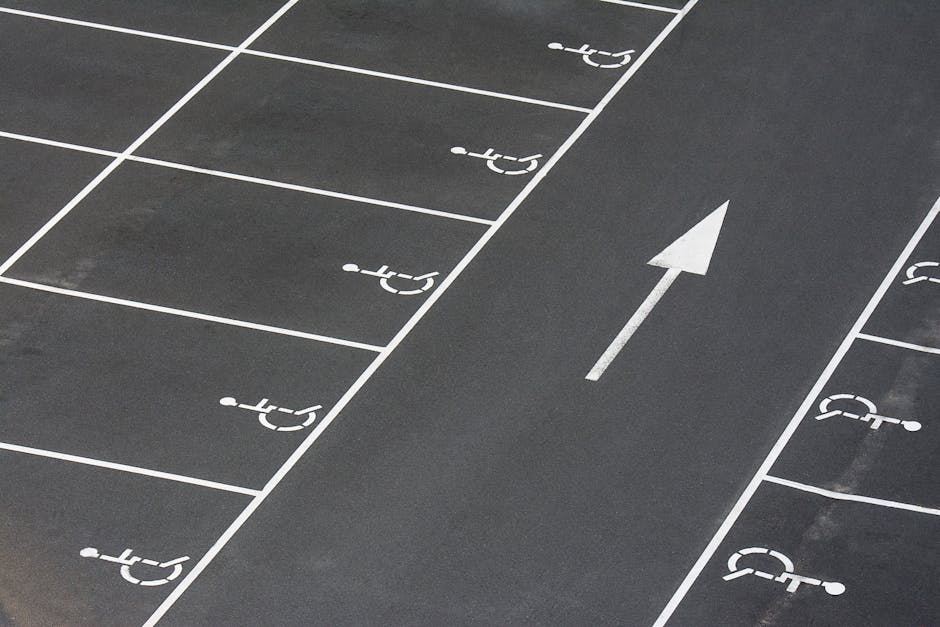 Close-up of a handicapped parking space symbol painted on an asphalt surface near the entrance of a property, with fading yellow lines and a worn wheelchair icon. The image shows the textured black asphalt with patches of lighter and darker areas, and the painted markings indicating designated parking for disabled individuals. The setting suggests the parking area is part of a home relocation or moving site, where a vehicle used by Man with Van Snaresbrook is parked nearby for furniture transport and loading. Visible around the edges are sections of the pavement, with no vehicles or furniture present in the frame, focusing solely on the parking space markings relevant to access and parking logistics for move-associated activities near Snaresbrook Crown Court.