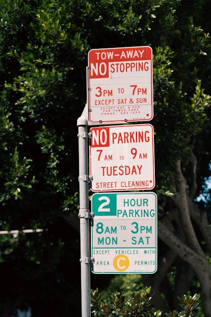 A tall metal post displaying multiple parking and towing regulation signs in a residential area, with a background of green trees and blue sky. The top sign is red and white, indicating a tow-away zone with no stopping from 3 pm to 7 pm, except on Saturdays and Sundays, with a phone number for inquiries. Below it, a second sign in red and white specifies no parking from 7 am to 9 am on Tuesdays for street cleaning. The third sign is blue and white, indicating a two-hour parking limit from 8 am to 3 pm, Monday through Saturday, with a note that it applies to vehicles with a C area permit. The signs are attached to the post with metal brackets, and the overall scene appears to be outdoors during daylight, illustrating typical parking restrictions in a suburban or urban street setting.