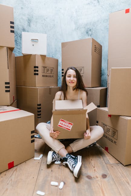 A young woman with long dark hair, wearing a beige top, beige pants, and black and white sneakers, is seated on a wooden floor inside a room filled with stacked cardboard moving boxes of various sizes, some with red 'Fragile' labels. She is holding an open cardboard box in her lap, with a slight smile on her face, surrounded by packing materials such as white packing peanuts scattered on the floor. The boxes are positioned against a textured light blue wall, indicating a home relocation process. Visible in the background are additional boxes stacked on top of each other, some sealed with packing tape. The scene depicts the packing and moving phase typical of professional removals services like those offered by Man with Van Snaresbrook, focusing on furniture transport, packing, and loading activities necessary for house removals and relocation support.