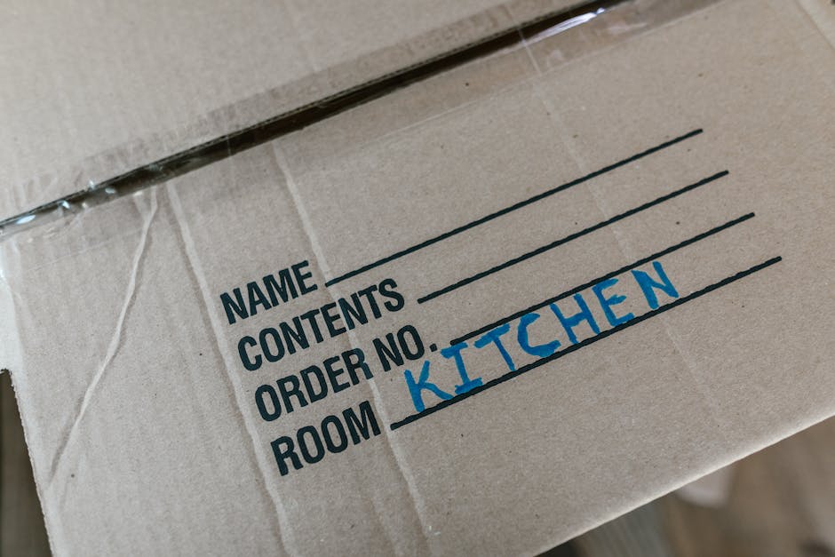 Close-up image of a cardboard moving box with printed sections for name, contents, order number, and room, with the word 'KITCHEN' handwritten in blue ink under the room section. The box is sealed with clear plastic packing tape and appears to be part of a home relocation or furniture transport process. The label indicates it contains kitchen items and is prepared for loading, fitting into a larger moving or packing operation managed by Man with Van Snaresbrook, a company offering removals services. The environment is neutral, and the focus is on the organized identification of contents for efficient packing and unpacking during house moves or relocation tasks.