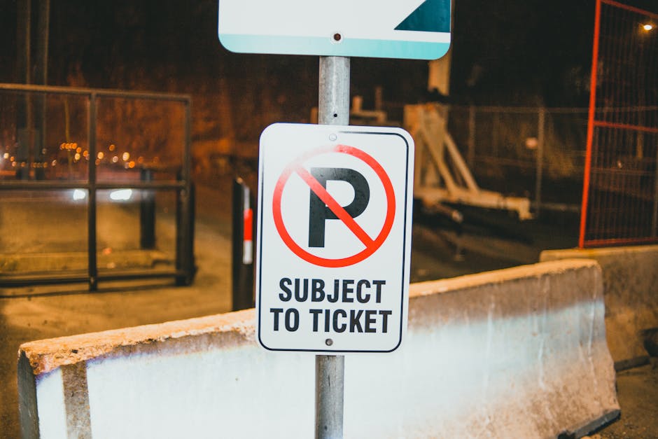 A white rectangular sign with a red circle and a diagonal line crossing out the letter 'P', indicating no parking, is mounted on a metal post positioned behind a low concrete barrier. Below the symbol, the sign reads 'SUBJECT TO TICKET' in black capital letters. The scene is outdoors at night, illuminated by ambient street lighting, with a metal fence or barrier to the left and background elements including construction equipment and a fence. The image depicts a parking restriction area near a property or building entrance, relevant to the logistical considerations of house removals and furniture transport. The setting aligns with the context of loading and unloading zones used in professional home relocation and moving services, with emphasis on clear signage to facilitate efficient moving operations. Man with Van Snaresbrook may utilize such signage to navigate parking rules during client moves.
