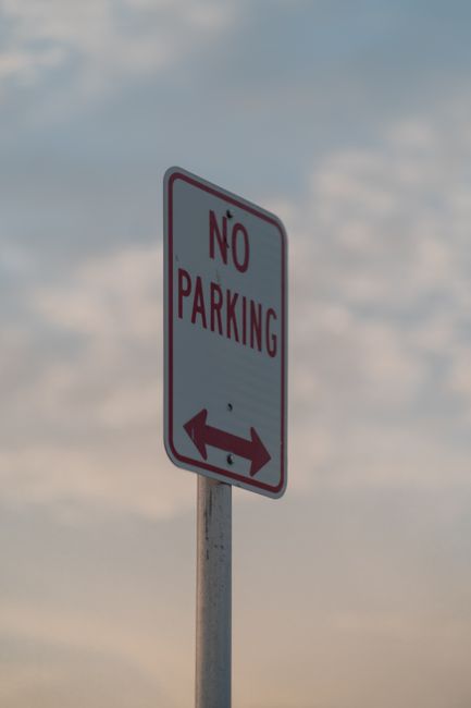 A white rectangular sign with a red circle and a diagonal line crossing out the letter 'P', indicating no parking, is mounted on a metal post positioned behind a low concrete barrier. Below the symbol, the sign reads 'SUBJECT TO TICKET' in black capital letters. The scene is outdoors at night, illuminated by ambient street lighting, with a metal fence or barrier to the left and background elements including construction equipment and a fence. The image depicts a parking restriction area near a property or building entrance, relevant to the logistical considerations of house removals and furniture transport. The setting aligns with the context of loading and unloading zones used in professional home relocation and moving services, with emphasis on clear signage to facilitate efficient moving operations. Man with Van Snaresbrook may utilize such signage to navigate parking rules during client moves.