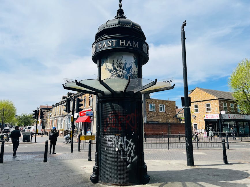 A tall, cylindrical street clock with a rounded top displaying the name 'East Ham' in white letters on a black background, situated on a busy pavement in an urban area. The clock is black with decorat