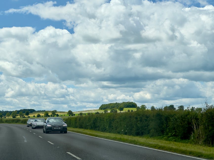 Photograph showing a rural UK road with three modern cars in a convoy, driving along a two-lane asphalt route bordered by green hedges and fields under a partly cloudy sky. In the background, there are rolling hills with patches of trees and grassy farmland. The scene is outdoors during daylight, with natural lighting illuminating the vehicles and landscape. This image may relate to home relocation or moving logistics, as it depicts transportation on the approach or departure from a property, aligning with moving and furniture transport services provided by Man with Van Snaresbrook, a company specializing in removals.