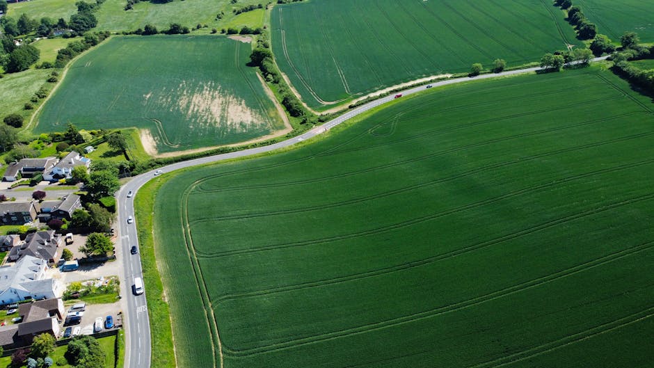 Photograph showing a rural UK road with three modern cars in a convoy, driving along a two-lane asphalt route bordered by green hedges and fields under a partly cloudy sky. In the background, there are rolling hills with patches of trees and grassy farmland. The scene is outdoors during daylight, with natural lighting illuminating the vehicles and landscape. This image may relate to home relocation or moving logistics, as it depicts transportation on the approach or departure from a property, aligning with moving and furniture transport services provided by Man with Van Snaresbrook, a company specializing in removals.