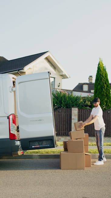 A man wearing a white T-shirt and grey pants is standing on the pavement beside a white moving van with its side door open, preparing to load cardboard boxes wrapped in brown packing tape into the vehicle. The boxes are stacked on the ground next to him, and he is holding one in his hands, ready to place it inside. The scene takes place outdoors in a residential area with a house that has a dark roof, a wooden fence, and tall green trees visible in the background. The sky appears clear, and the lighting suggests daylight, indicating a home relocation or furniture transport process managed by Man with Van Snaresbrook, focused on packing and loading for a house removal.